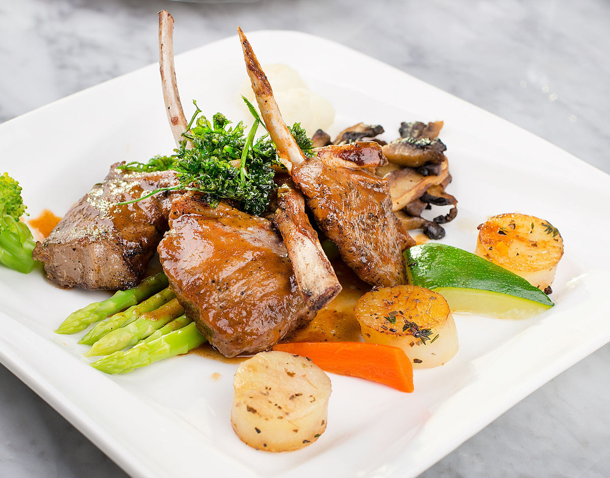 A plated gourmet dish featuring lamb chops with bone-in pieces, mushrooms, green asparagus, scallops, broccoli, and a lime wedge on a white square plate against a light gray marble background.
cooked wth Saffronia Threads