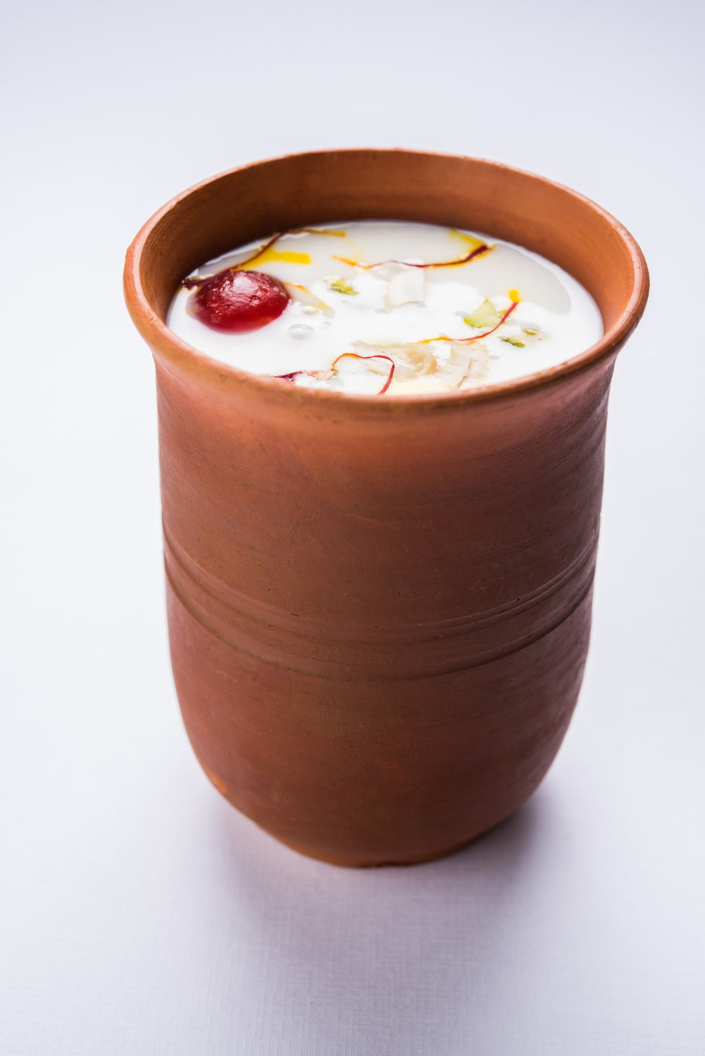 A terracotta clay cup filled with a white beverage, garnished with a red berry, white coconut pieces, and yellow and red threads, set against a clean white background.