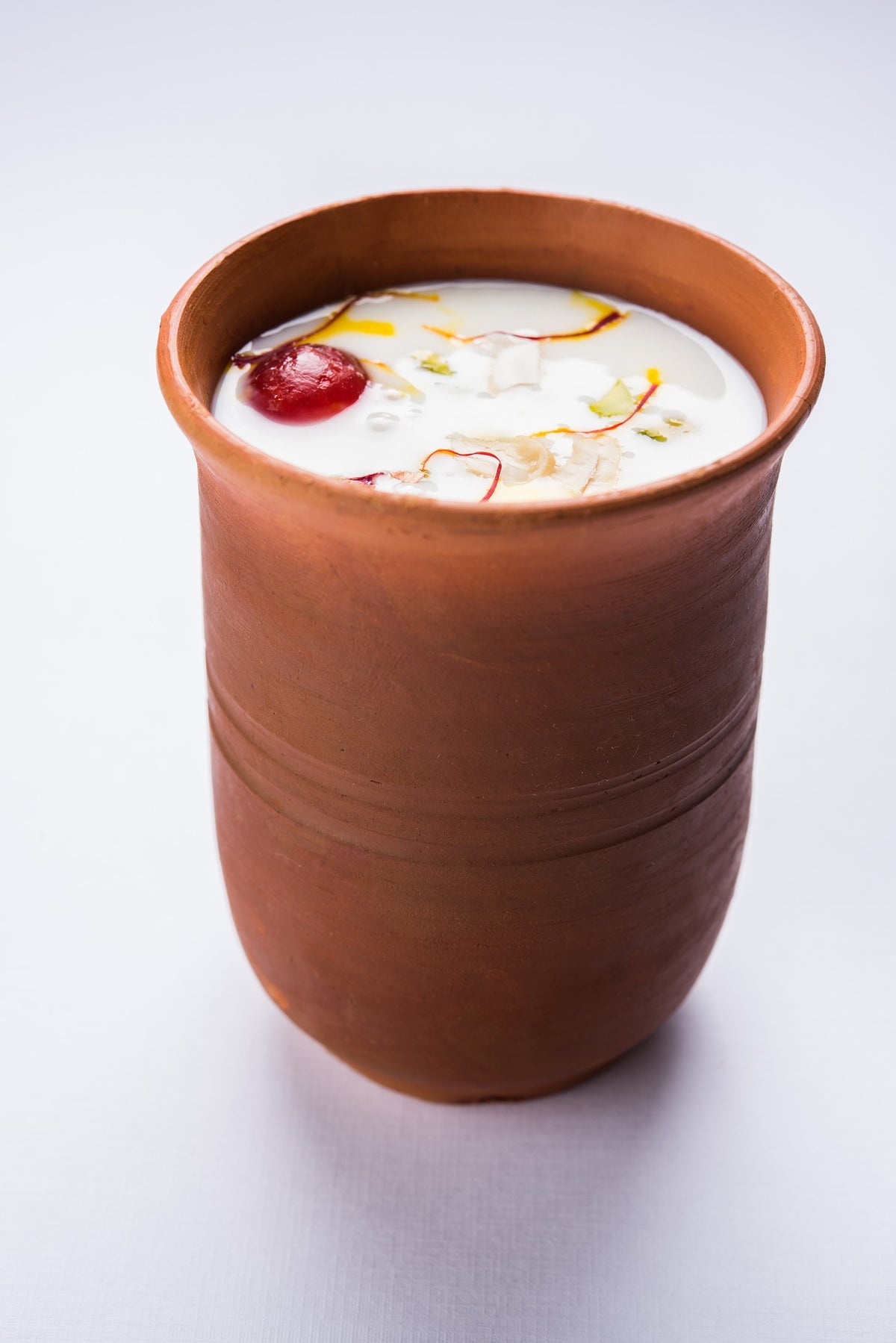 A terracotta clay cup filled with a white beverage, garnished with a red berry, white coconut pieces, and yellow and red threads, set against a clean white background.